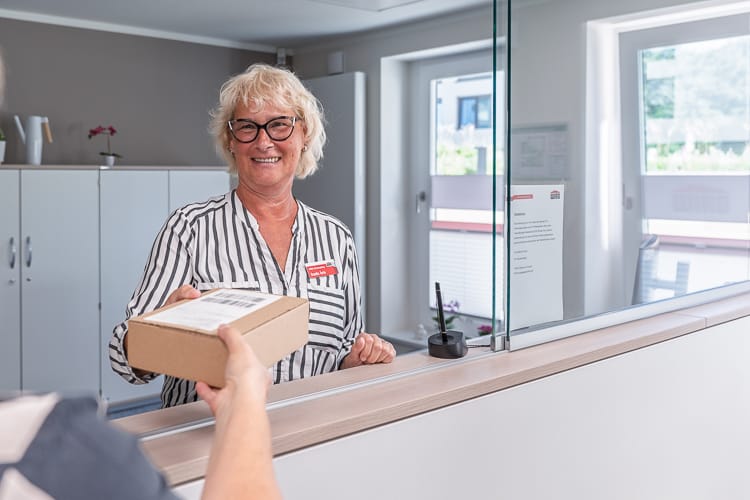 Freundliche, lächelnde Mitarbeiterin im Hausempfang mit gestreifter Bluse übergibt hinter einem Schalter aus Holz und Glas ein kleines Paket an eine Person im Vordergrund.
