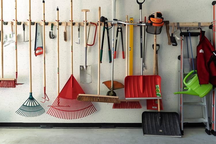 Eine aufgeräumte Werkzeugwand in einer Garage, an der Besen, Spaten, Rechen, Schaufeln und Gartengeräte übersichtlich an einer Holzleiste mit Haken hängen.