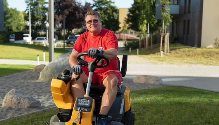 Ein Hausmeister auf einem modernen Aufsitzmäher. Er mäht den Rasen in einer modernen Wohnanlage. Er blickt freundlich in die Kamera.