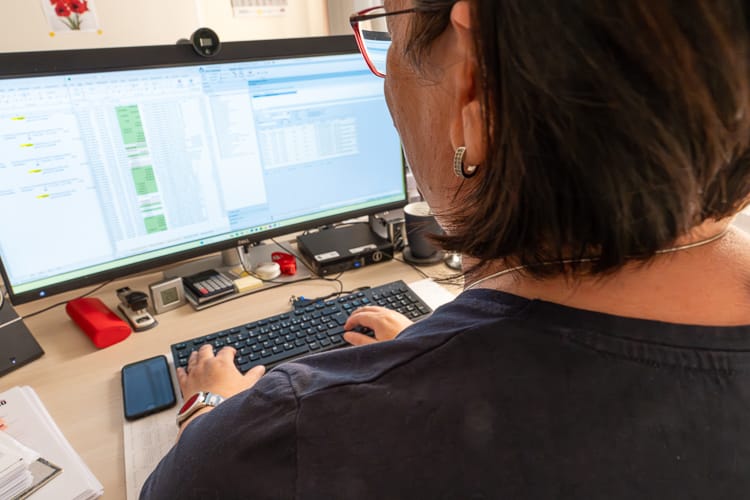 Blick über die Schulter einer Mitarbeiterin auf einen großen Monitor mit vielen Tabellen.