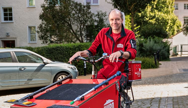 Lächelnder Reinigungs-in WIRO-Arbeitskleidung fährt auf einem roten Lastenfahrrad auf einer gepflasterten Straße in Rostock.