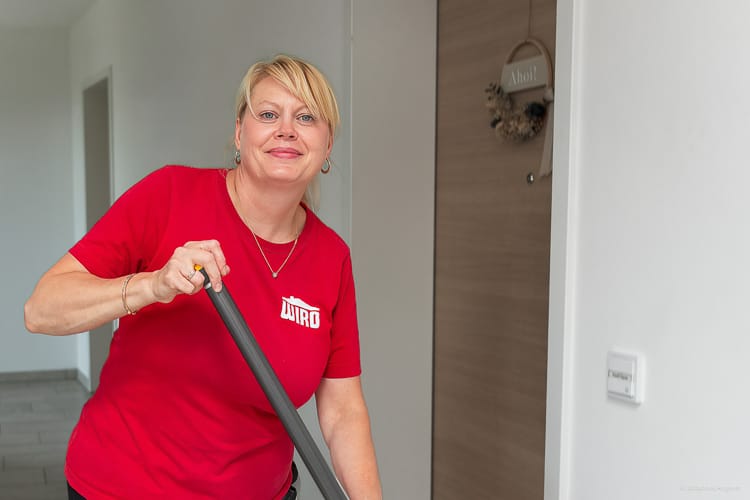Lächelnde WIRO-Mitarbeiterin in rotem T-Shirt mit Logo, die mit einem Wischmopp einen Flur eines Wohnhauses reinigt. 