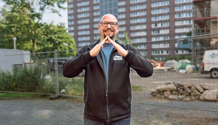 Ein Sozialarbeiter der WIRO steht im Freien und formt mit seinen Händen das WIRO-Dach. Im Hintergrund sieht man eine große Wohnanlage. 