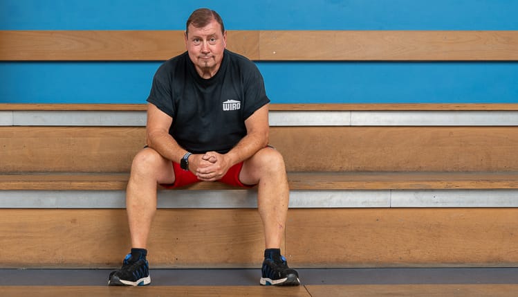 Ein Hallenwart der WIRO sitzt im schwarzen T-Shirt und kurzen roten Hosen auf einer hölzernen Tribüne vor einer blauen Wand.