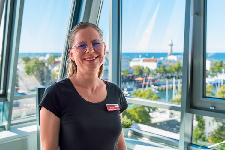Lächelnde Hotelmanagerin in einem schwarzen T-Shirt mit Namensschild. Sie steht vor einer großen Glasfront mit Blick auf Warnemünde und die Ostsee.