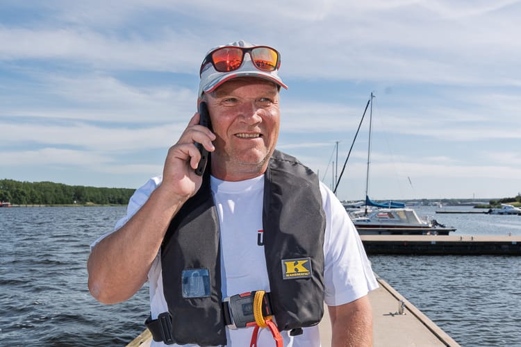 Lässig lächelnder Hafenmeister der WIRO in weißem T-Shirt, dunkler Rettungsweste und Baseball-Cap. Er telefoniert an einem sonnigen Tag mit seinem Handy auf einem Steg vor Segelbooten.
