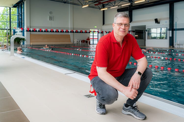 lächelnder Mitarbeiter der Schwimmhalle in rotem Polo-Shirt und Jeans, der vor dem gefliesten Beckenrand in der Schwimmhalle kniet. Im Hintergrund ist das große Schwimmbecken mit Bahnen zu sehen.