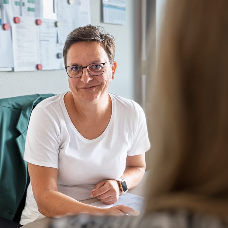 Lächelnde Azubiverantwortliche der WIRO sitzt im Büro am Schreibtisch. Sie trägt ein weißes T-Shirt und eine Brille, im Hintergrund ist eine Pinnwand mit Notizen zu sehen.
