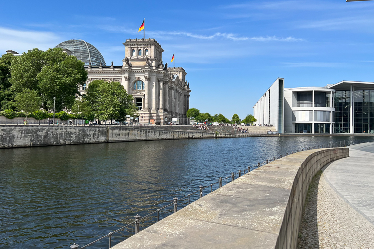 Außenansicht des Reichstagsgebäudes in Berlin mit der Reichstagskuppel und wehenden deutschen Flaggen, aufgenommen von der Uferpromenade der Spree bei Sonnenschein. Rechts ist ein modernes Regierungsgebäude zu sehen.
