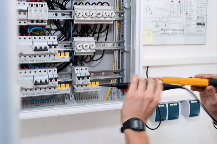 Nahaufnahme der Hände eines Elektrikers, der mit einem gelben Prüfgerät Messungen an den Sicherungen und Komponenten eines offenen Stromverteilerkastens  vornimmt.