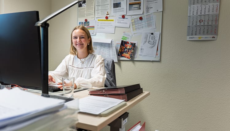 Lächelnde WIRO-Auszubildende sitzt an ihrem aufgeräumten Schreibtisch vor einem Computer. Im Hintergrund hängt eine Pinnwand mit Notizen und ein Kalender.