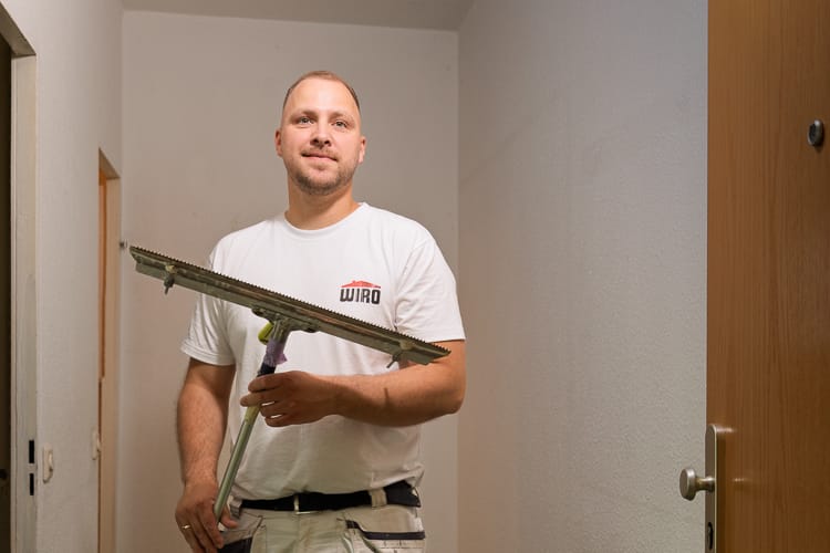 Lächelnder Maler und Lackierer der WIRO in weißem T-Shirt mit Logo. Er steht in einem unrenovierten Flur und hält eine Werkzeug zum Auftragen von Kleber oder Spachtelmasse, eine Zahntraufel, in die Kamera.