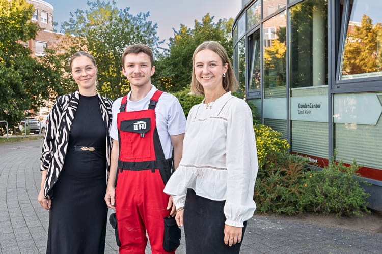 Drei junge WIRO-Mitarbeiter stehen lächelnd vor dem KundenCenter Groß Klein. Der Mann trägt eine rote Latzhose, die Frauen tragen elegante Business-Kleidung.