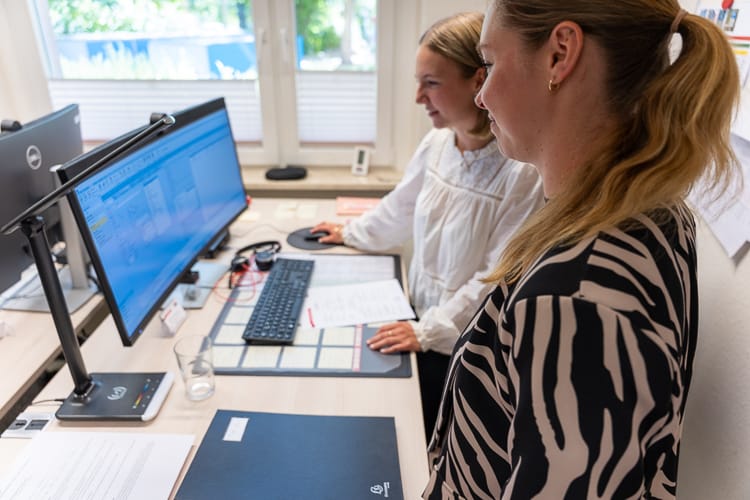 wei lächelnde WIRO-Mitarbeiterinnen, eine Azubi und ihre Ausbilderin, stehen gemeinsam vor einem Computer am Schreibtisch und besprechen Inhalte auf dem Bildschirm im Büro.