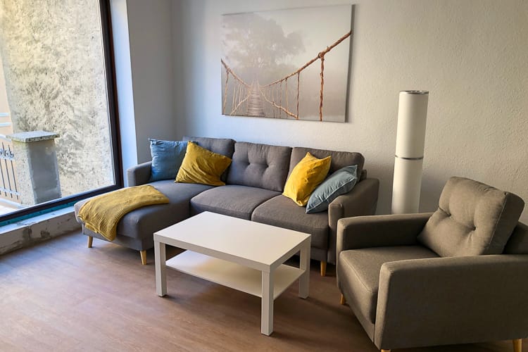 Das helle, modern eingerichtete Wohnzimmer unsere Azubi-WG in Waren. Mit grauem Ecksofa, grauem Sessel und weißem Couchtisch.