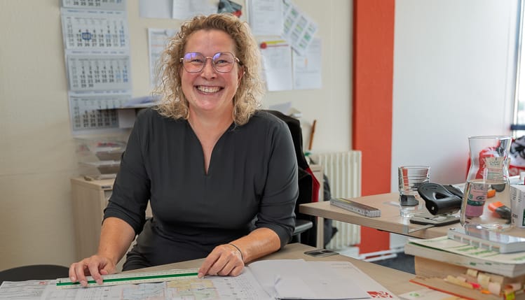 Lächelnde Bauingenieurin der WIRO mit Brille sitzt am Schreibtisch in ihrem Büro. Sie arbeitet an Bauplänen und hält ein Lineal in der Hand.