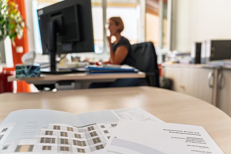 Nahaufnahme eines Bauprojektdokuments mit dem Bauplan eines Mehrfamilienhauses auf einem Schreibtisch. Im unscharfen Hintergrund arbeitet eine Mitarbeiterin am Computer.