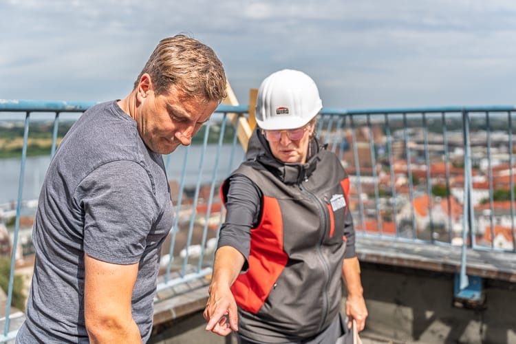 Zwei WIRO-Mitarbeiter bei der Besprechung von Baudetails auf einem Dach, mit Blick auf eine Stadt. Die Frau trägt einen weißen Schutzhelm mit WIRO-Logo.
