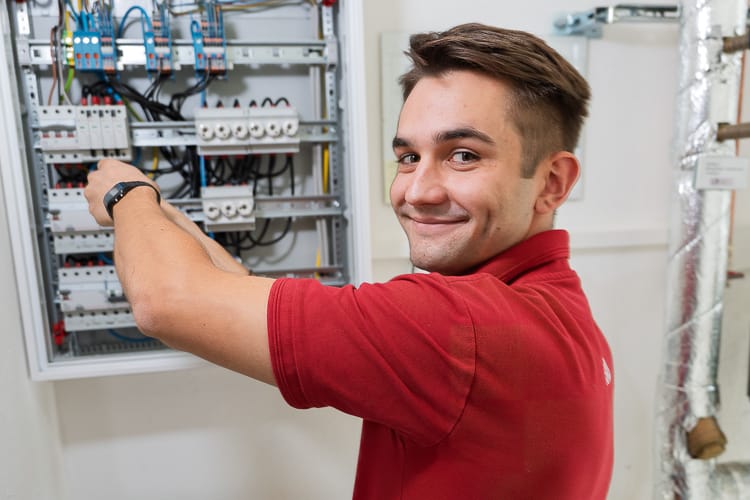 Elektriker schraubt etwas an einem Schaltkasten. Er blickt über die Schulter freundlich in die Kamera.
