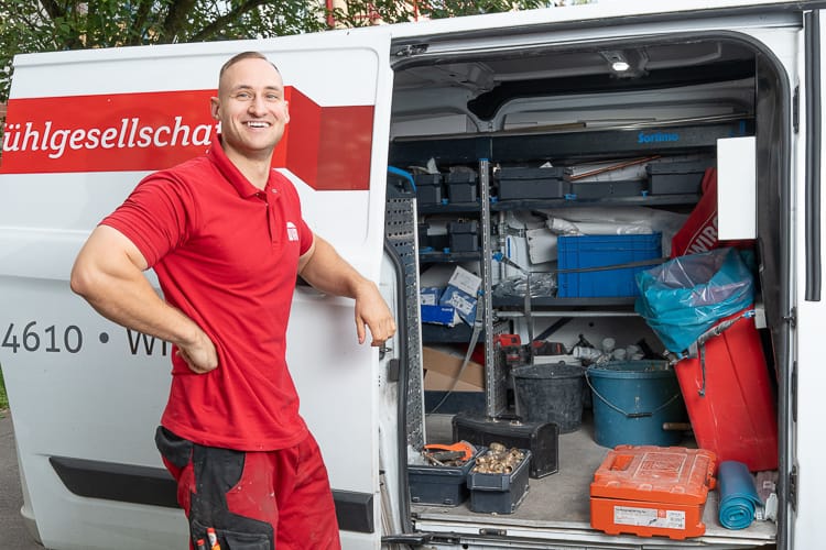 Installateur steht vor seinem Transporter neben der geöffneten Seitentüre. Im Auto sieht man SHK-Werkzeug.