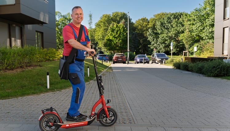 Ein WIRO-Hausmeister steht vor einer Wohnanlage auf einem E-Scooter. Er blickt freundlich in die Kamera. 
