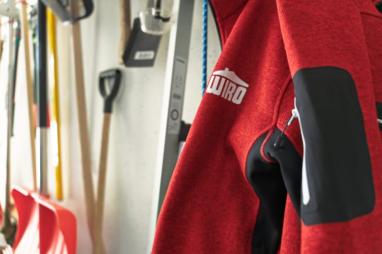 Im Vordergrund eine aufgehängte WIRO-Jacke, unscharf dahinter sieht man an einer Wand Stielwerkzeuge. 