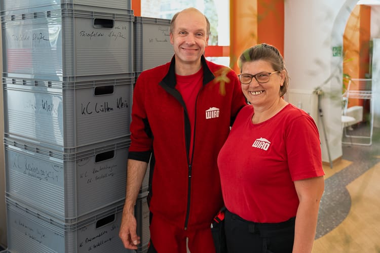 Zwei Mitarbeiter der Gebäudereinigung stehen vor einem Stapel grauer Kisten mit Reinigungsmaterial. Der Mann und die Frau blicken freundlich lächelnd in die Kamera. 