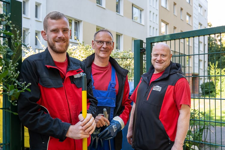 Drei junge WIRO-Mitarbeiter stehen lächelnd vor dem KundenCenter Groß Klein. Der Mann trägt eine rote Latzhose, die Frauen tragen elegante Business-Kleidung.