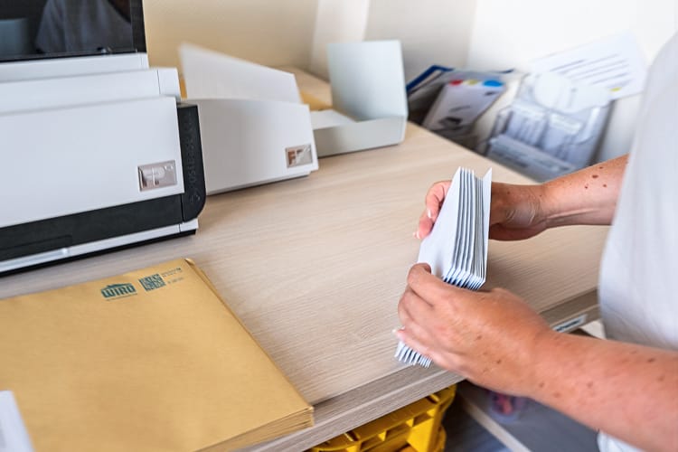 Hände, die ein Bündel Briefe halten. Dahinter eine Frankiermaschine.