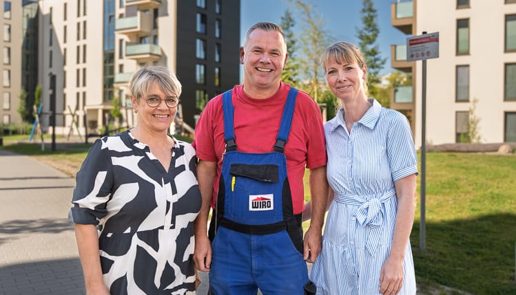 3 WIRO-Mitarbeiter, ein Mann und zwei Frauen, stehen in einer Wohnanlage und lächeln freundlich in die Kamera