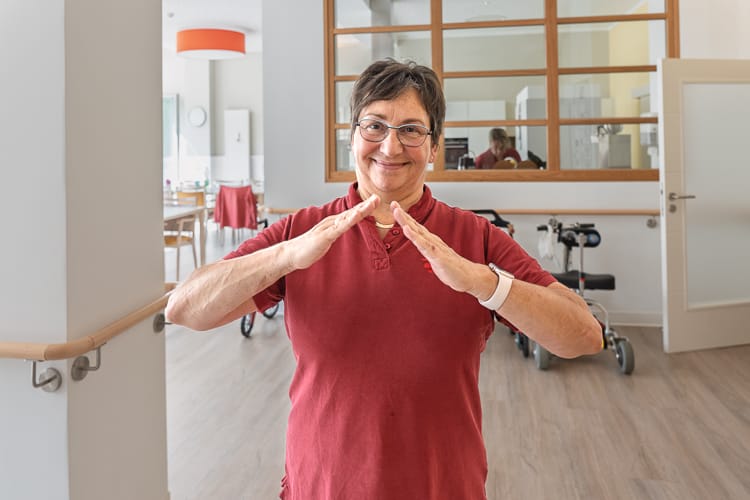 Lächelnde Mitarbeiterin der Tagespflege der WIRO. Sie trägt ein rotes Polo-Shirt und formt in einem hellen Gemeinschaftsraum mit ihren Händen die Dachgeste des WIRO-Logos.