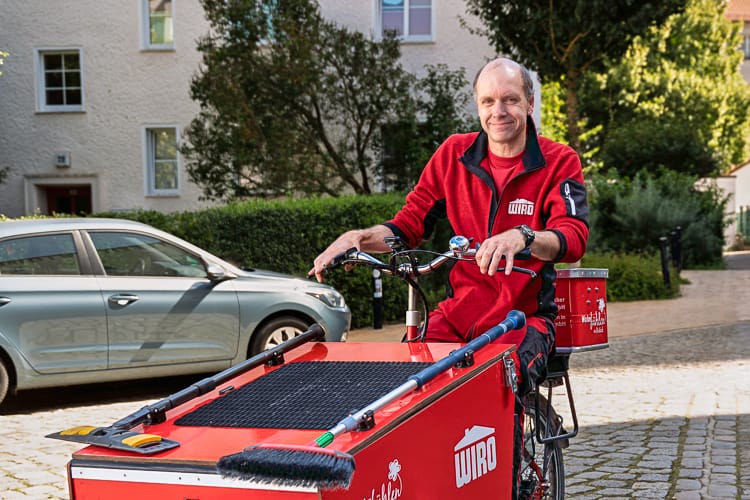 Lächelnder Reinigungsmitarbeiter WIRO-Arbeitskleidung fährt auf einem roten Lastenfahrrad auf einer gepflasterten Straße in Rostock.