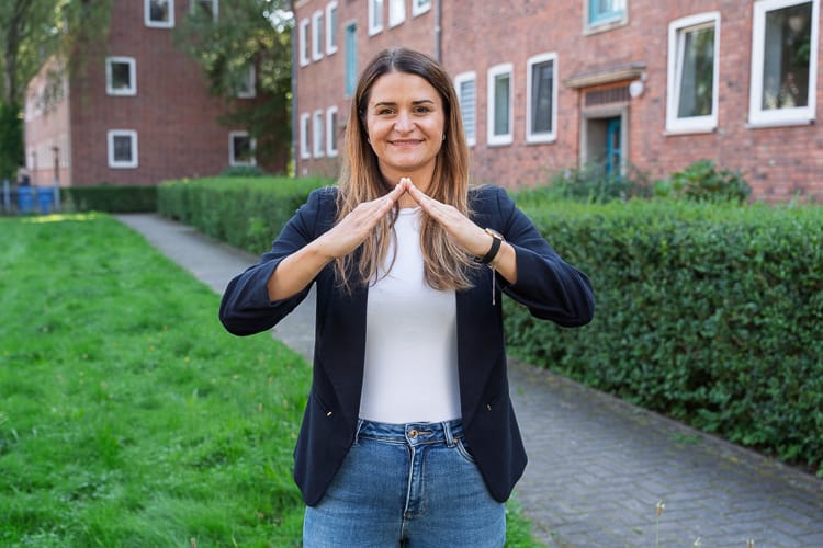 Lächelnde Mitarbeiterin der WEG-Abteilung der WIRO steht im Freien vor einem roten Backsteingebäude. Sie trägt einen dunklen Blazer und Jeans und formt mit ihren Händen das Dach des WIRO-Logos.